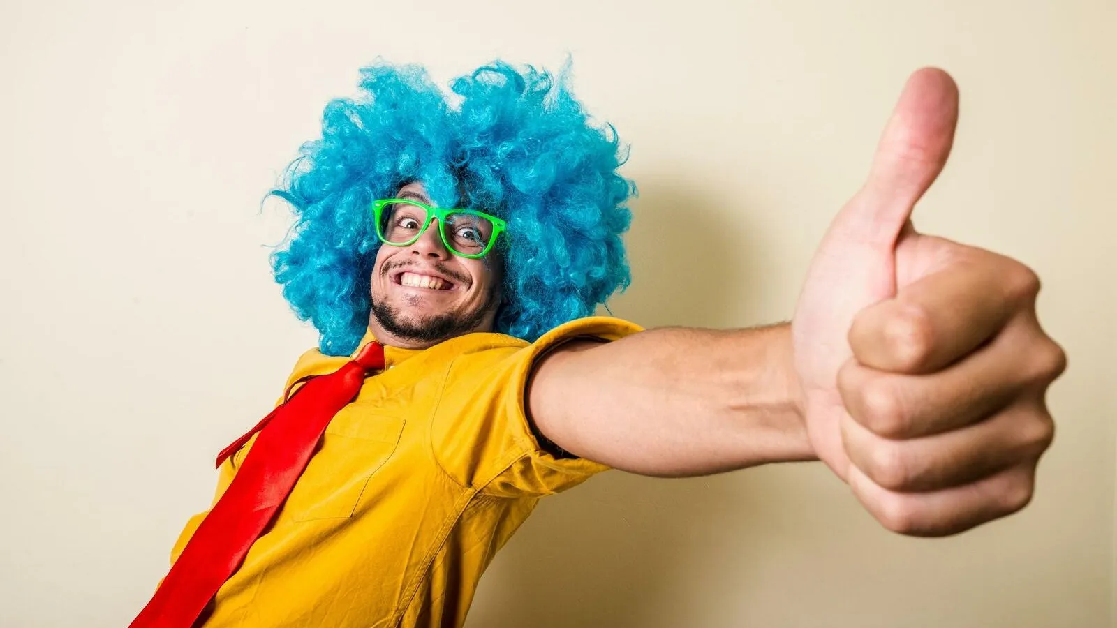 Ein Mann mit einer blauen Perücke, einer grünen Brille, einem gelben Shirt und einer roten Krawatte zeigt den Daumen hoch.