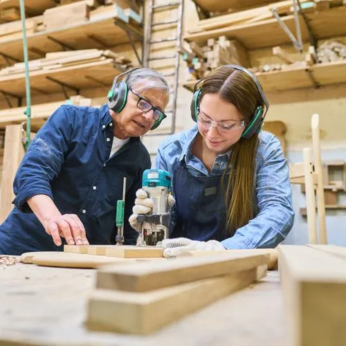 Eine junge Frau fräst ein Stück Holz aus und ihr Kollege schaut dabei zu.