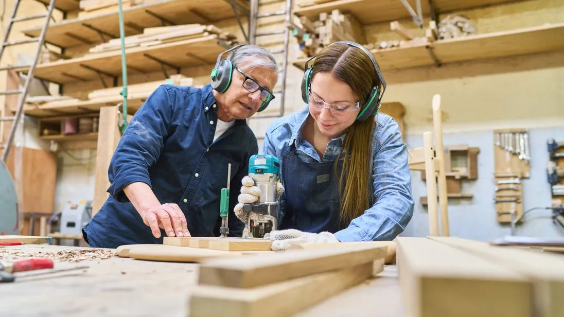 Eine junge Frau fräst ein Stück Holz aus und ihr Kollege schaut dabei zu.