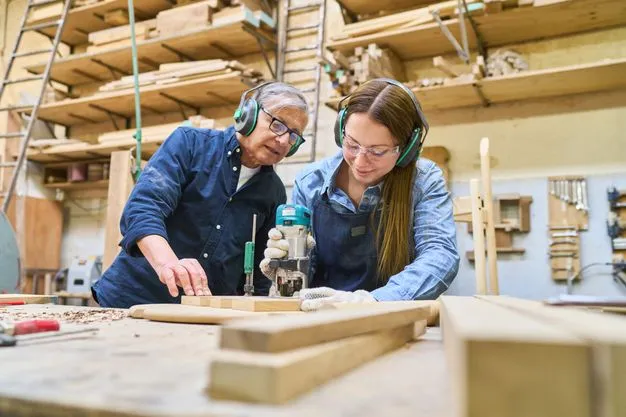 Eine junge Frau fräst ein Stück Holz aus und ihr Kollege schaut dabei zu.