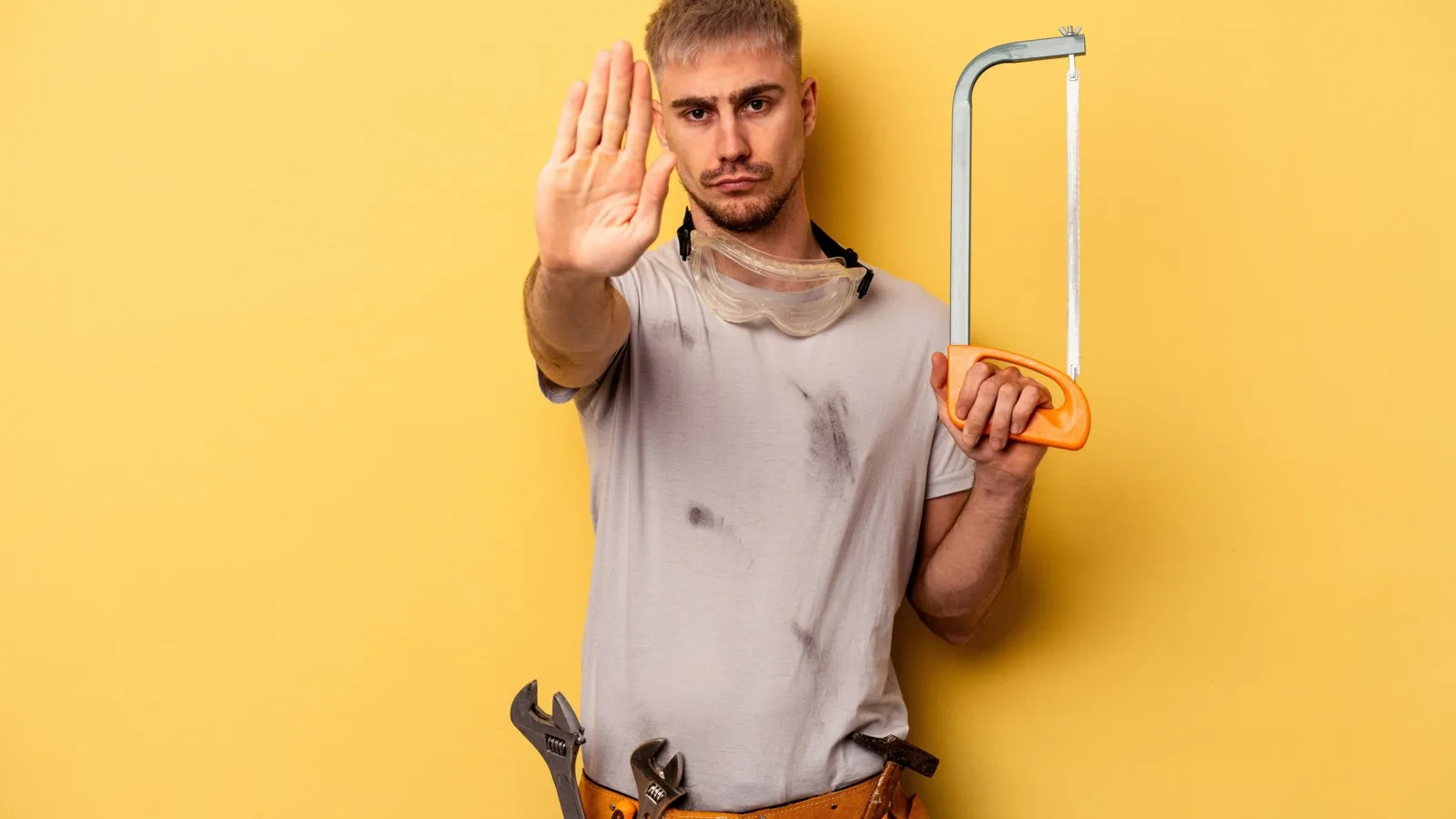 Ein junger Handwerker steht mit ausgestreckter Hand, welche "Stopp" signalisieren soll, vor einem gelben Hintergrund. In seiner anderen Hand hält er eine Bügelsäge.