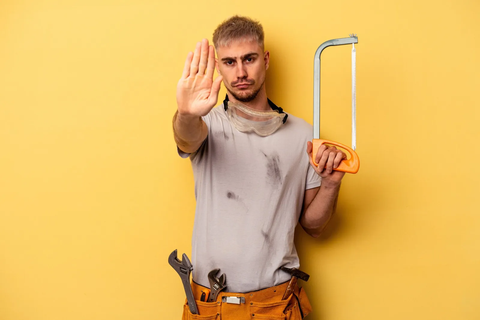 Ein junger Handwerker steht mit ausgestreckter Hand, welche "Stopp" signalisieren soll, vor einem gelben Hintergrund. In seiner anderen Hand hält er eine Bügelsäge.