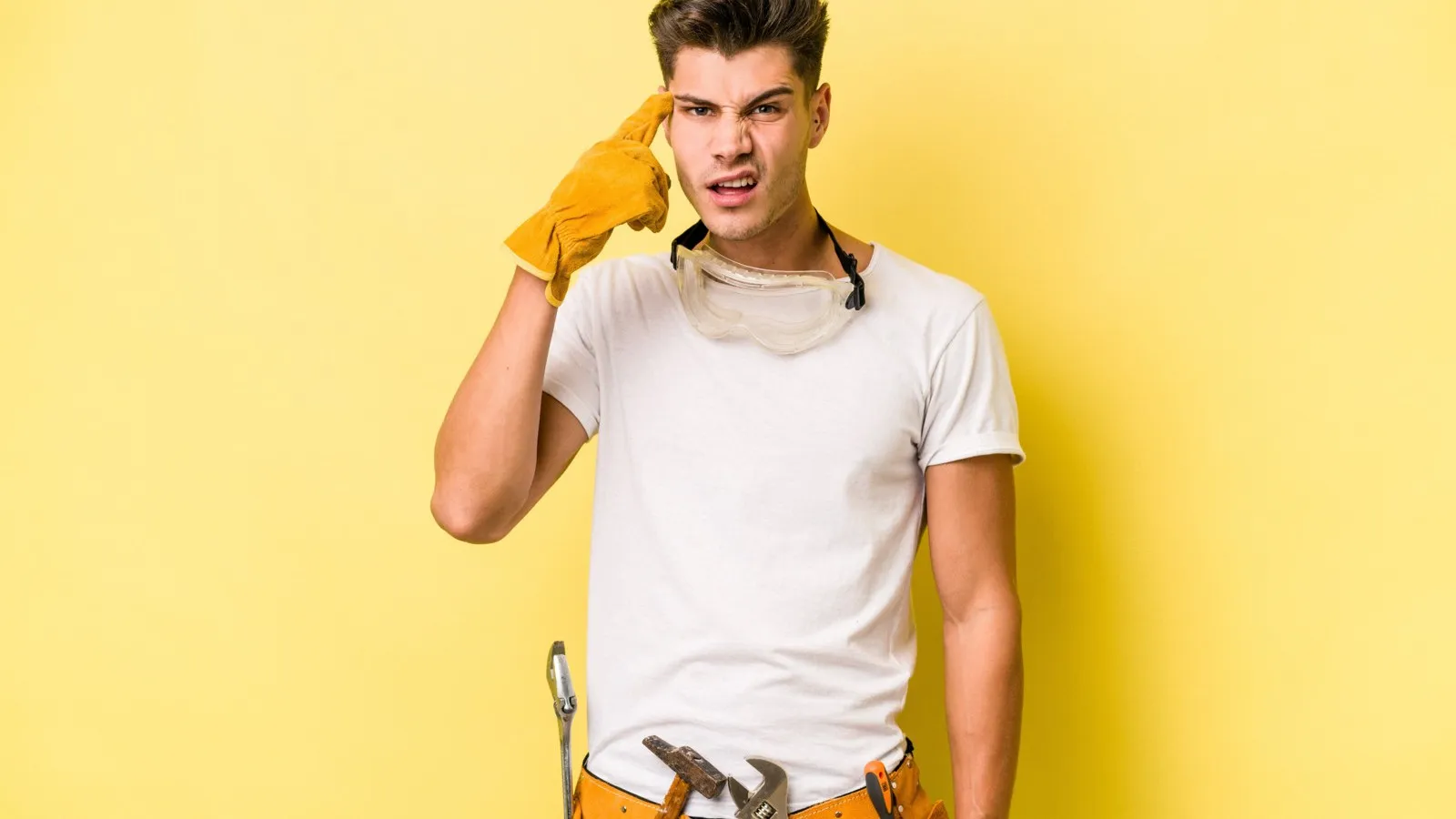 Ein junger Handwerker mit gelben Handschuhen steht vor einem hellgelben Hintergrund. Er zeigt mit dem Finger auf seine Schläfe und verzieht das Gesicht aus Unverständnis.