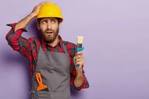 Nervous construction worker holds paint brush, touches head and looks aside, wears safety hardhat, dressed in apron, puzzled with having much work, poses on purple wall with blank copy space