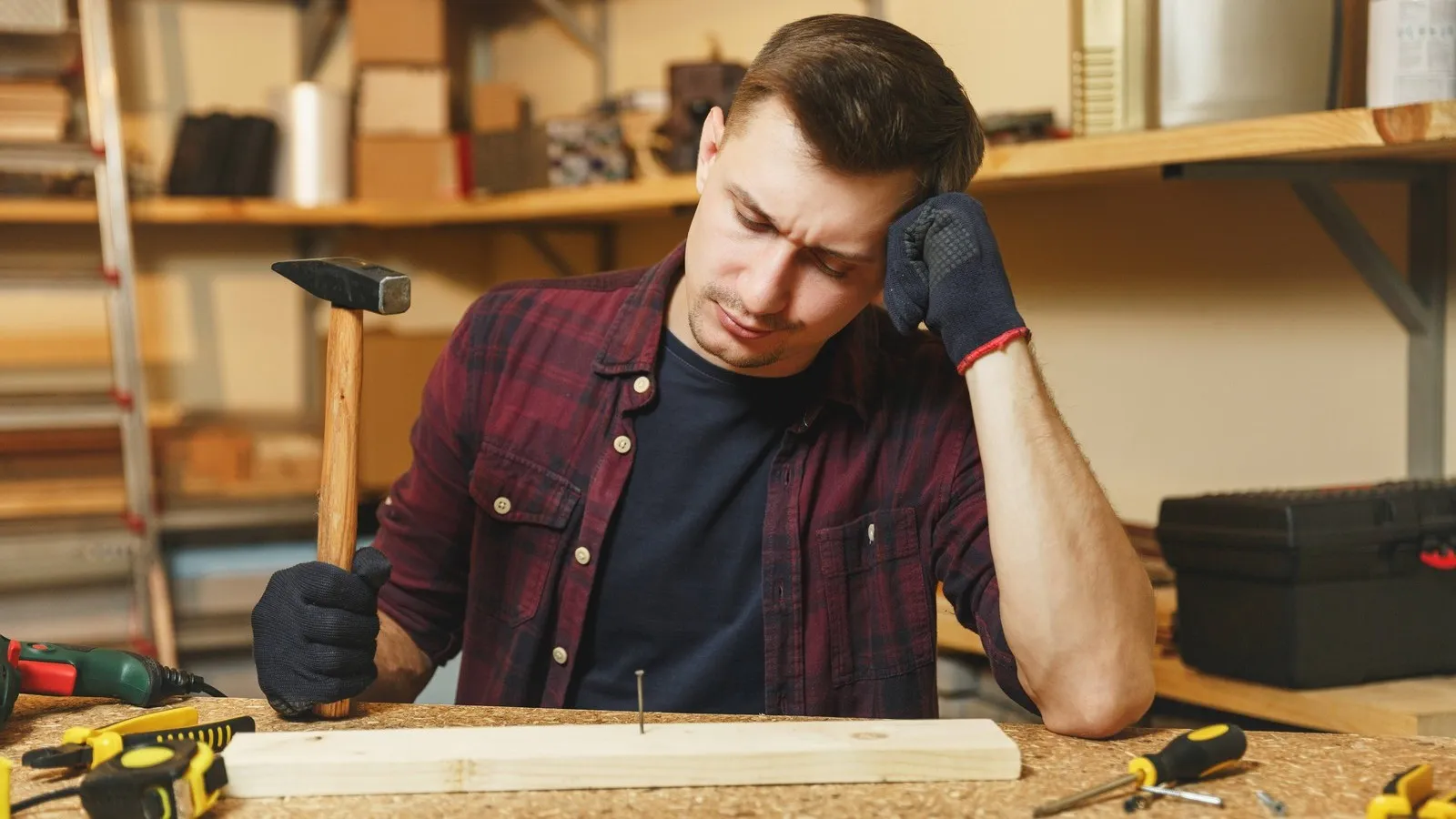 Müder und frustrierter Handwerker in einer Schreinerei beim Einschlagen von Nägeln in ein Holzstück an einer Werkbank, umgeben von Werkzeugen.