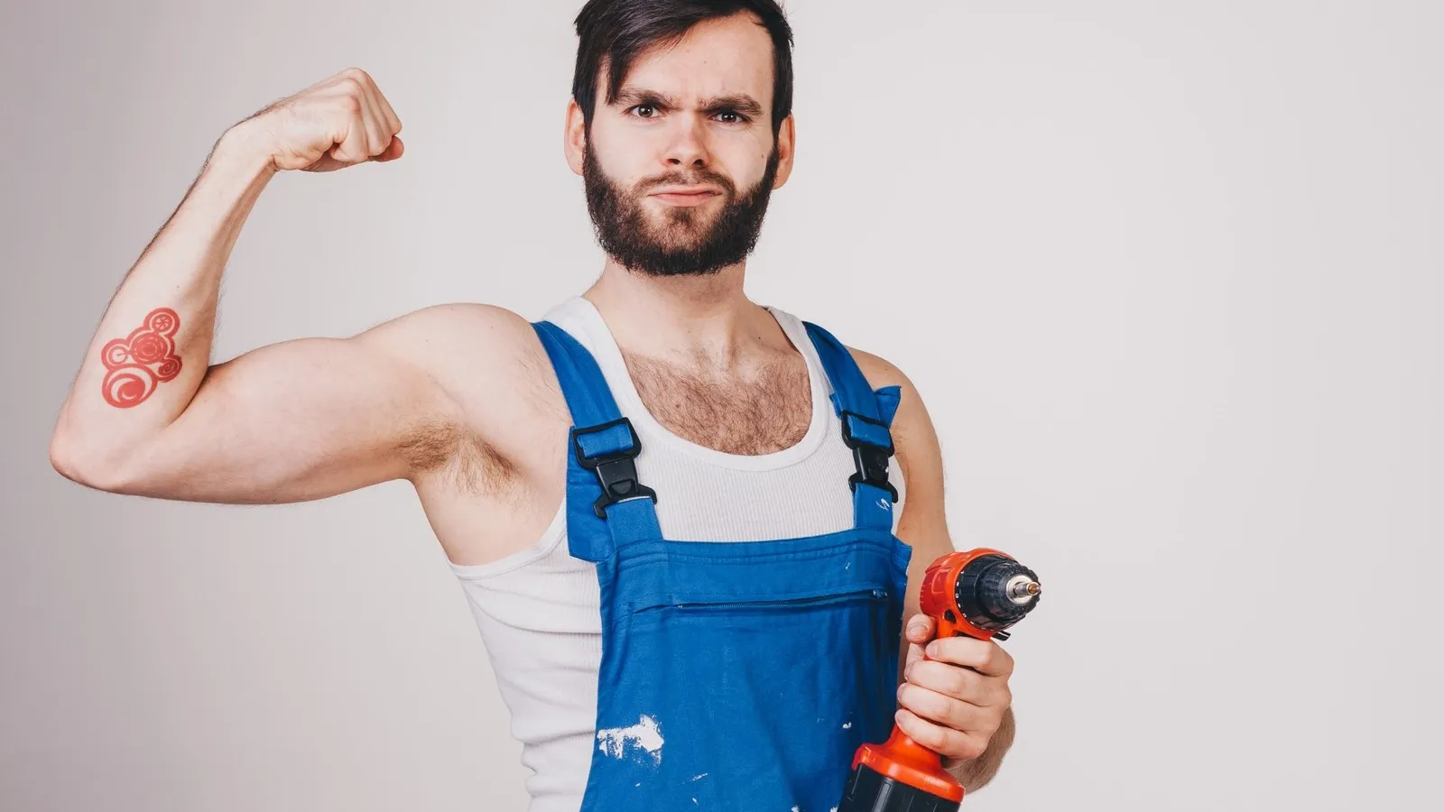 Handwerker in Arbeitskleidung spielt mit Muskeln und zeigt Bohrmaschine.