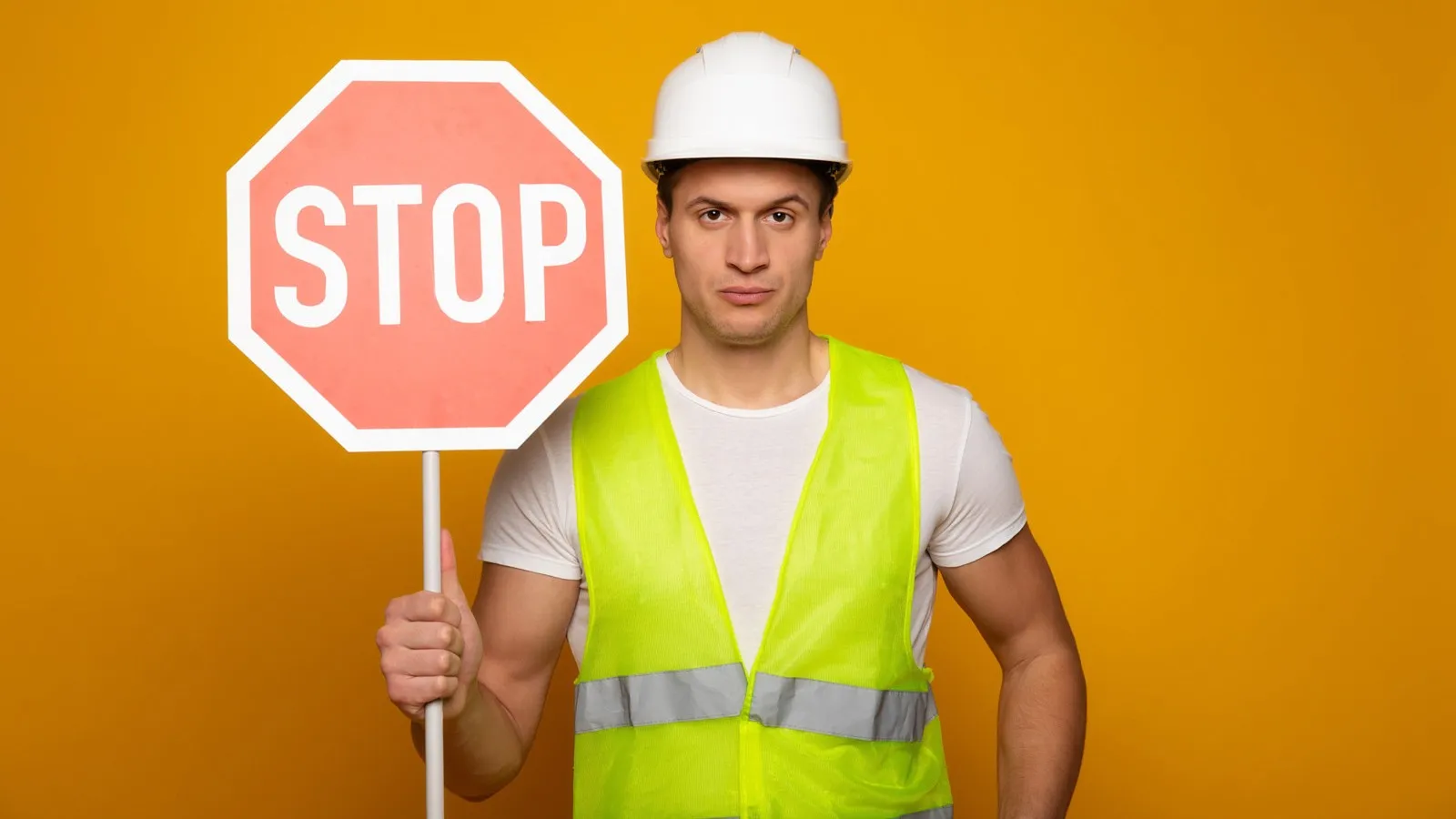 Ein Mann mit einem weißen Schutzhelm und einer gelben Warnweste steht vor einem orangenen Hintergrund und hält ein Stoppschild hoch.