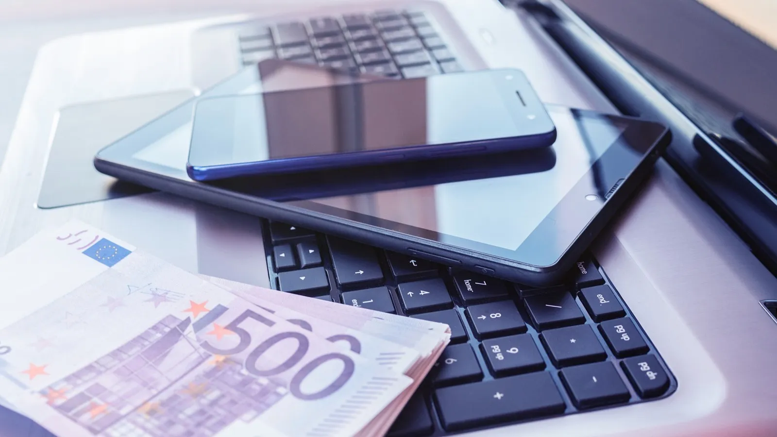 TEin Tablet liegt auf einer Laptoptastatur, daneben liegen 500-Euro Scheine in einem Bündel.