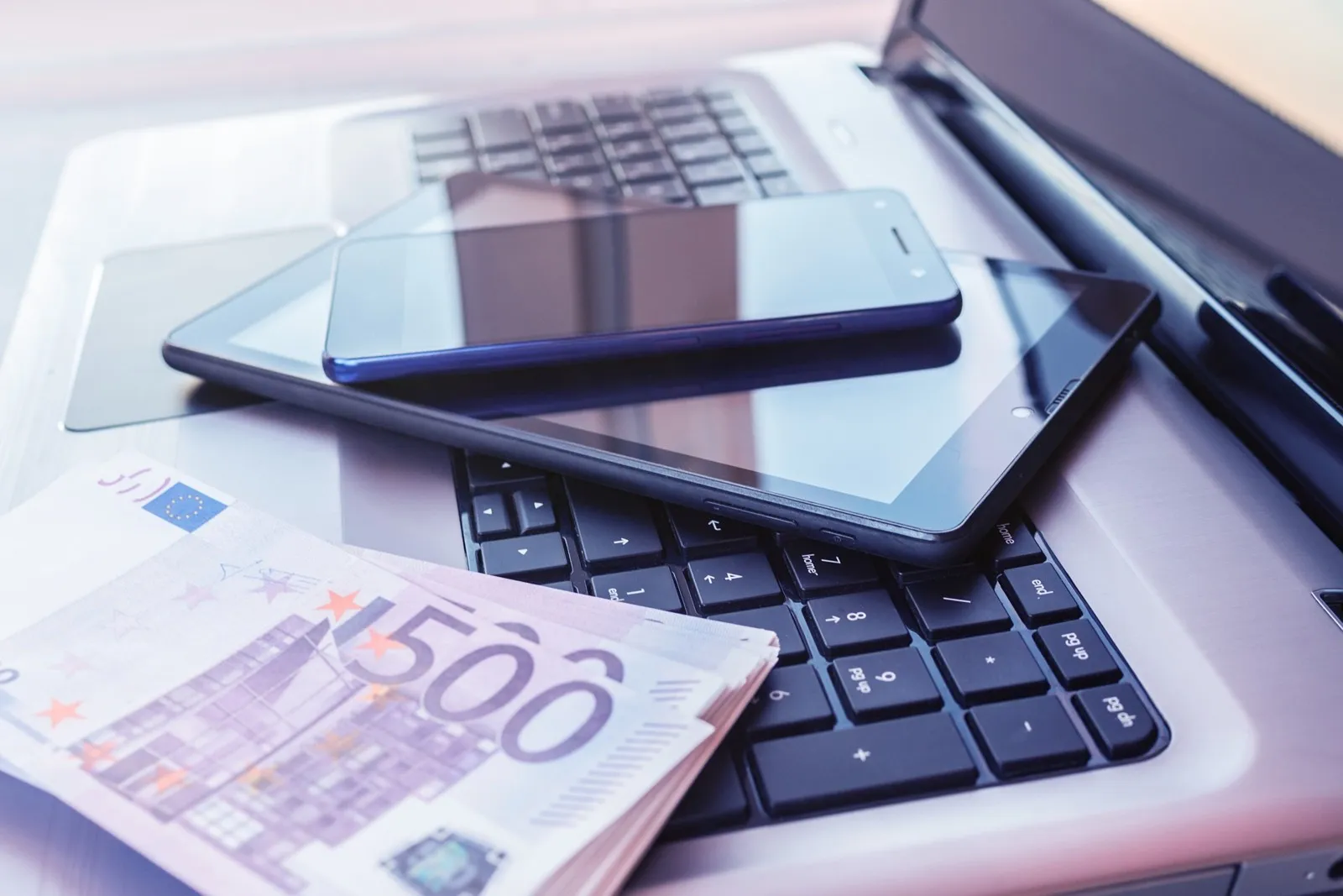 TEin Tablet liegt auf einer Laptoptastatur, daneben liegen 500-Euro Scheine in einem Bündel.