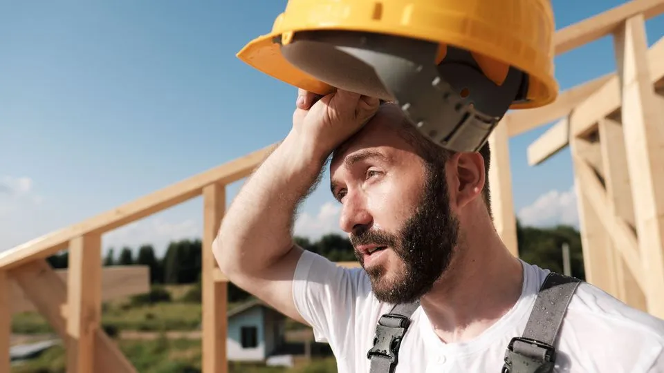 Ein Handwerker wischt sich mit dem Handrücken den Schweiß von der Stirn, er hat einen gelben Bauhelm in der Hand.