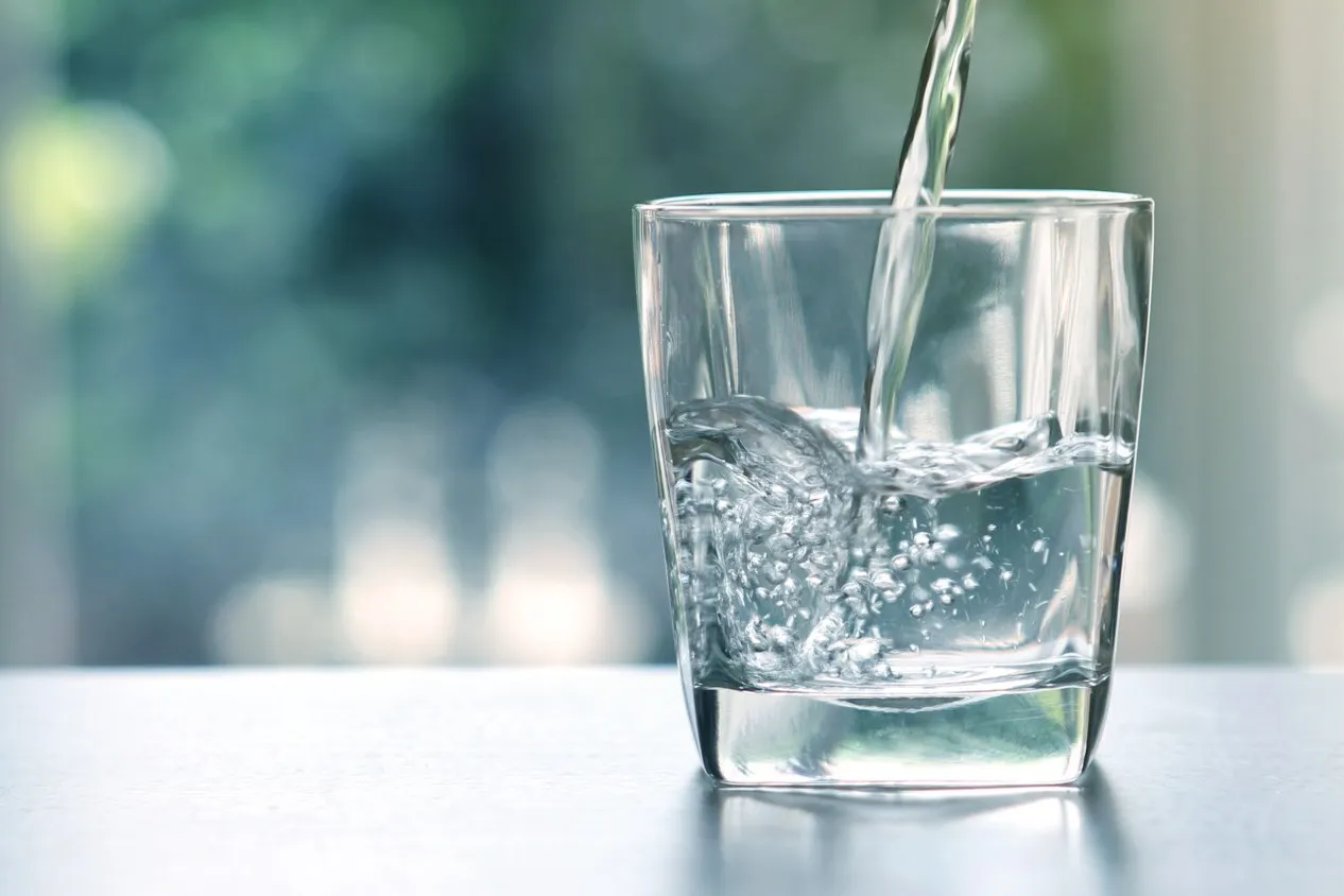 Ein Glas Wasser steht auf einem Tisch. Es wird mehr Wasser ins Glas gegossen.