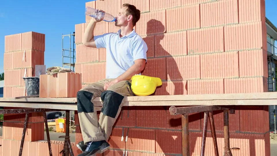 Handwerker sitzt auf einem Gerüst auf der Baustelle in der Sonne und trinkt etwas.