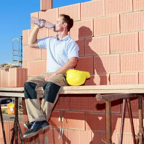 Handwerker sitzt auf einem Gerüst auf der Baustelle in der Sonne und trinkt etwas.