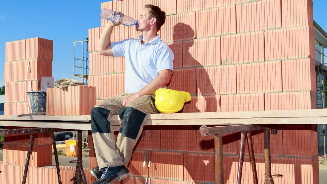 Handwerker sitzt auf einem Gerüst auf der Baustelle in der Sonne und trinkt etwas.