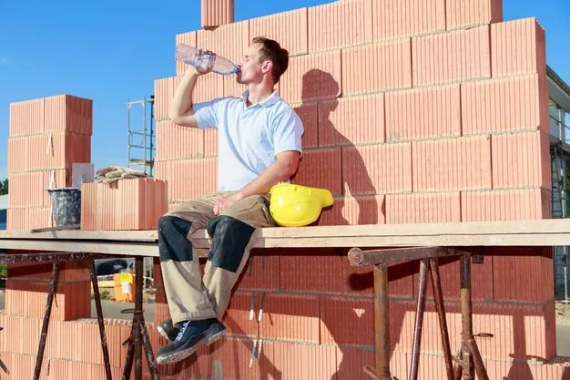 Handwerker sitzt auf einem Gerüst auf der Baustelle in der Sonne und trinkt etwas.
