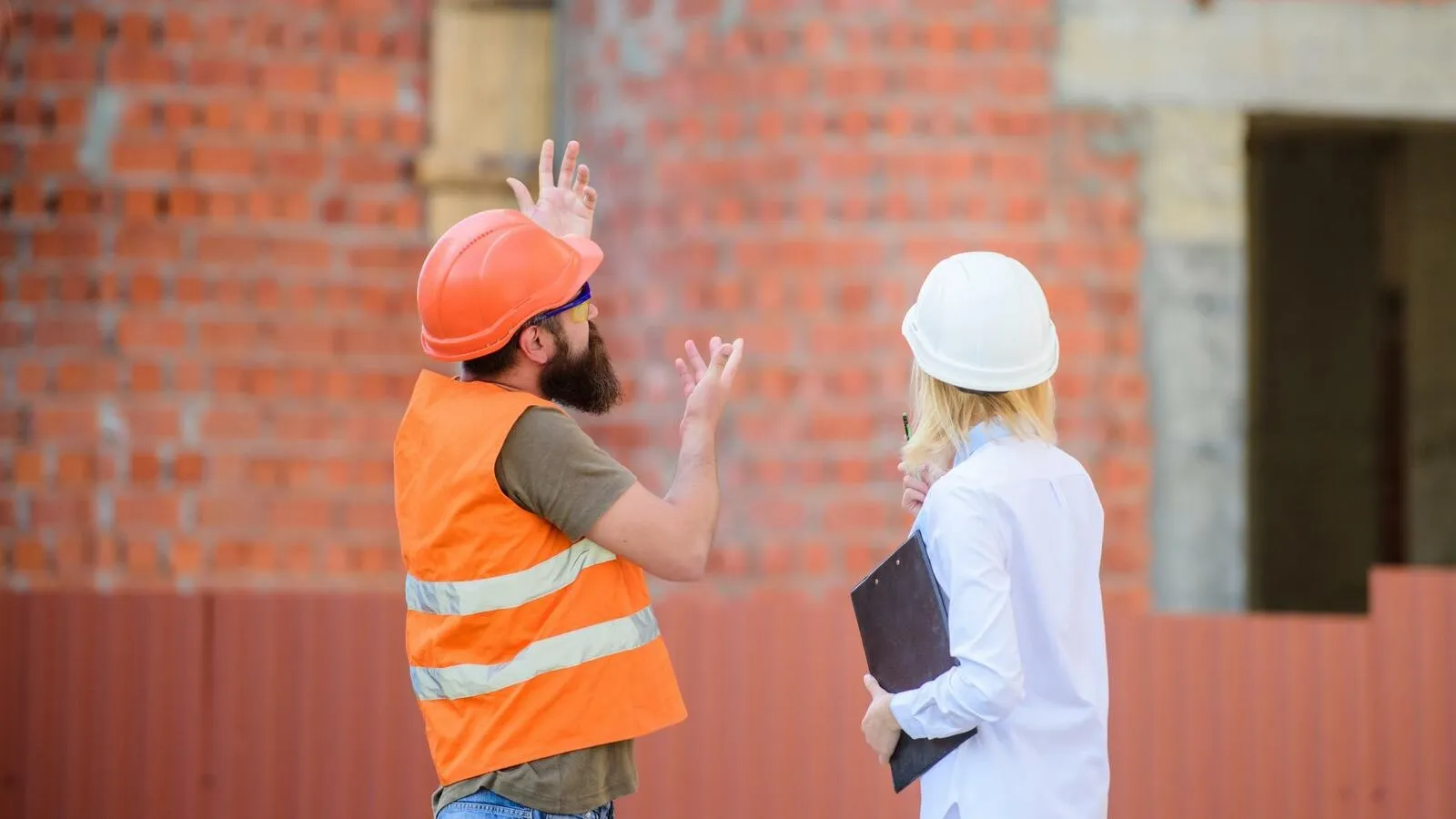 Ein Bauarbeiter mit orangenem Schutzhelm und Warnweste argumentiert mit einer weiß gekleideten Frau mit einem weißen Schutzhelm.