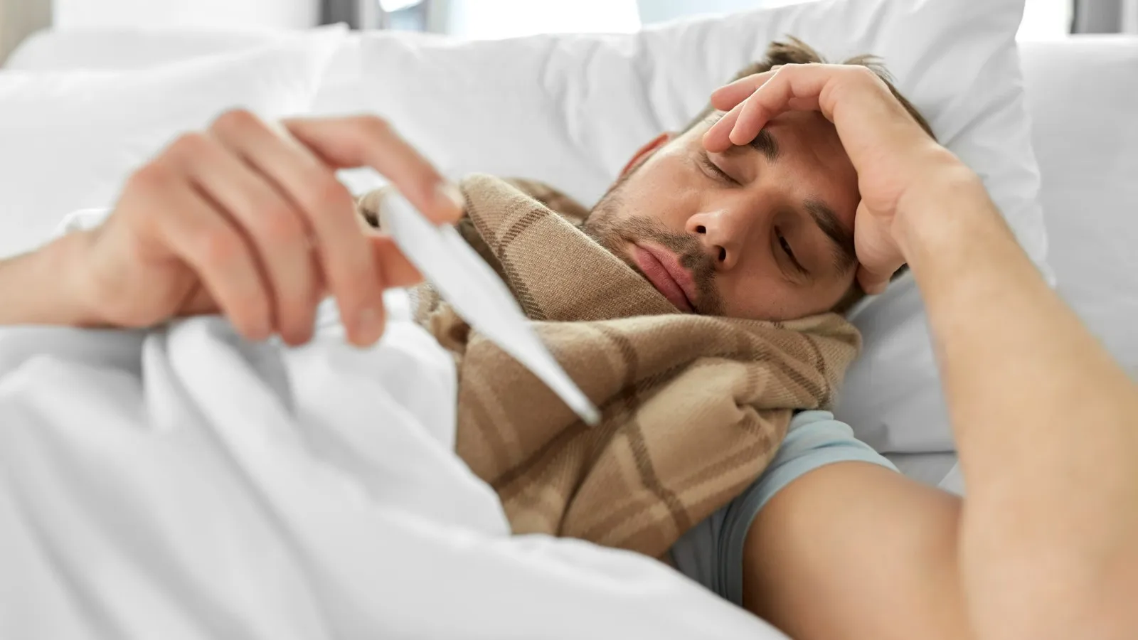 Ein kranker Mann liegt im Bett und schaut auf sein Thermometer.