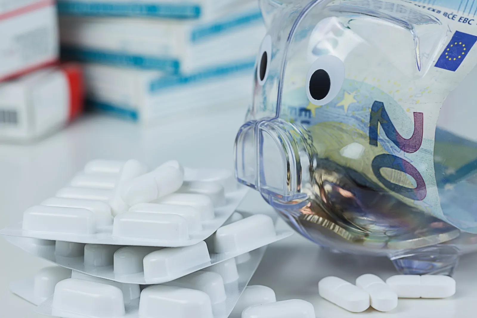 Ein 20 Euro-Schein steckt in einem durchsichtigen Sparschwein, daneben liegen mehrere Blister und lose Tabletten.