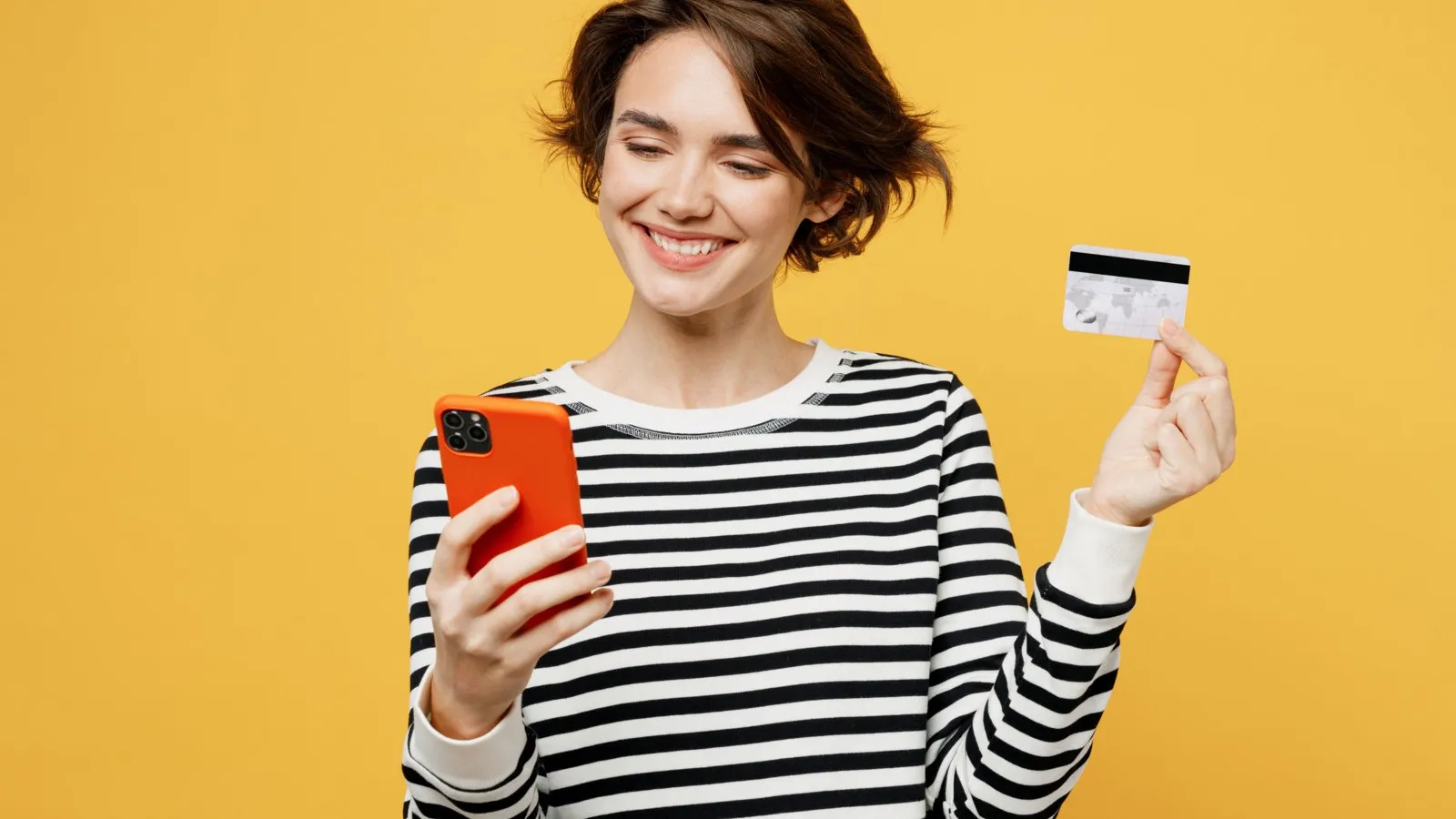 Eine Frau mit einem Handy in der Hand steht vor einem gelben Hintergrund. In der anderen Hand hält sie eine Kreditkarte hoch.