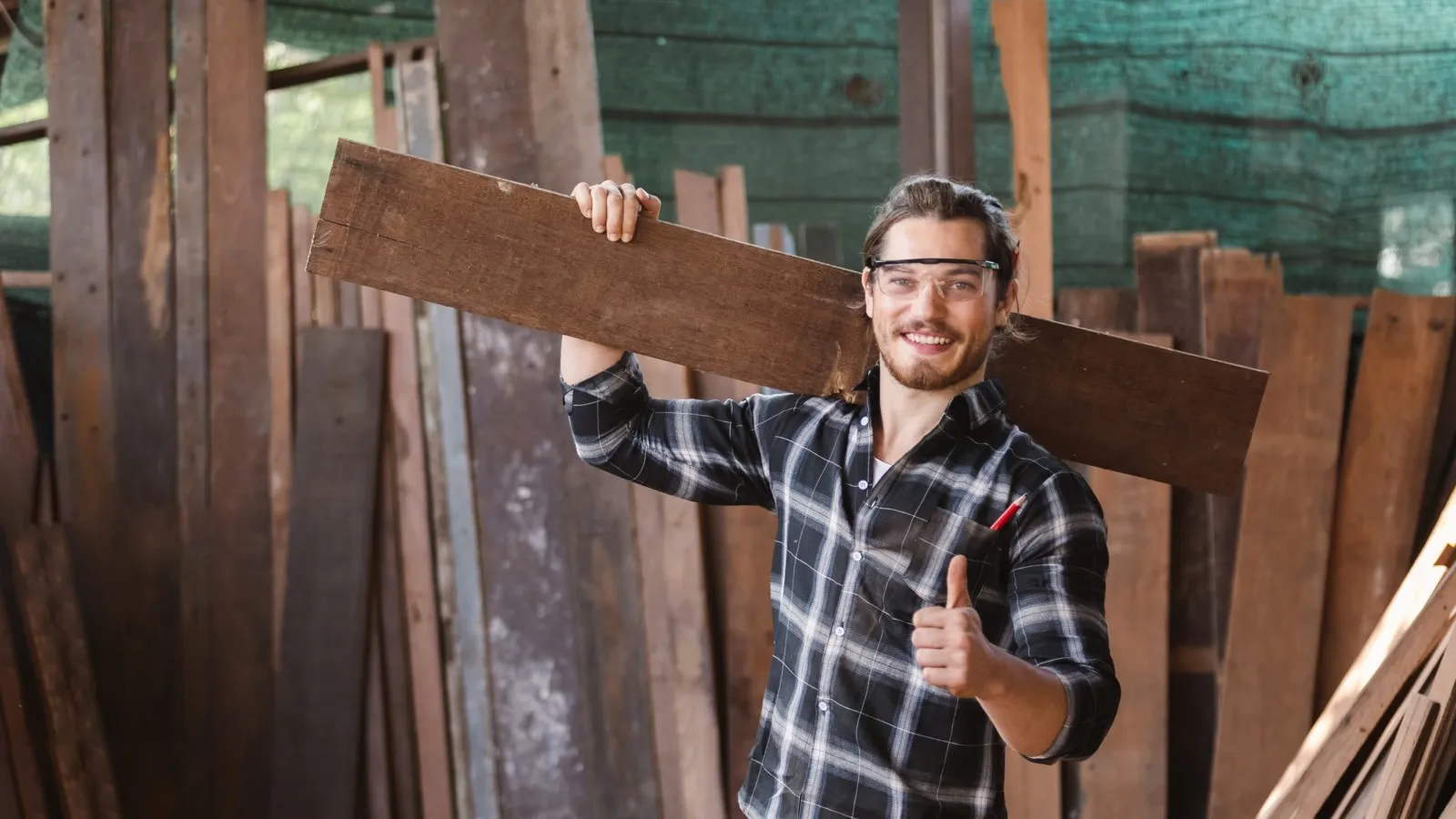 Ein Handwerker trägt ein Holzbrett auf seiner Schulter, lächelt und zeigt den Daumen hoch.