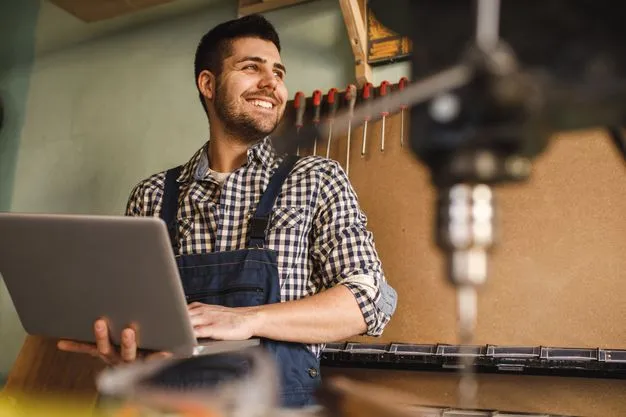 Tischler steht mit einem Laptop in seiner Werkstatt und lächelt.