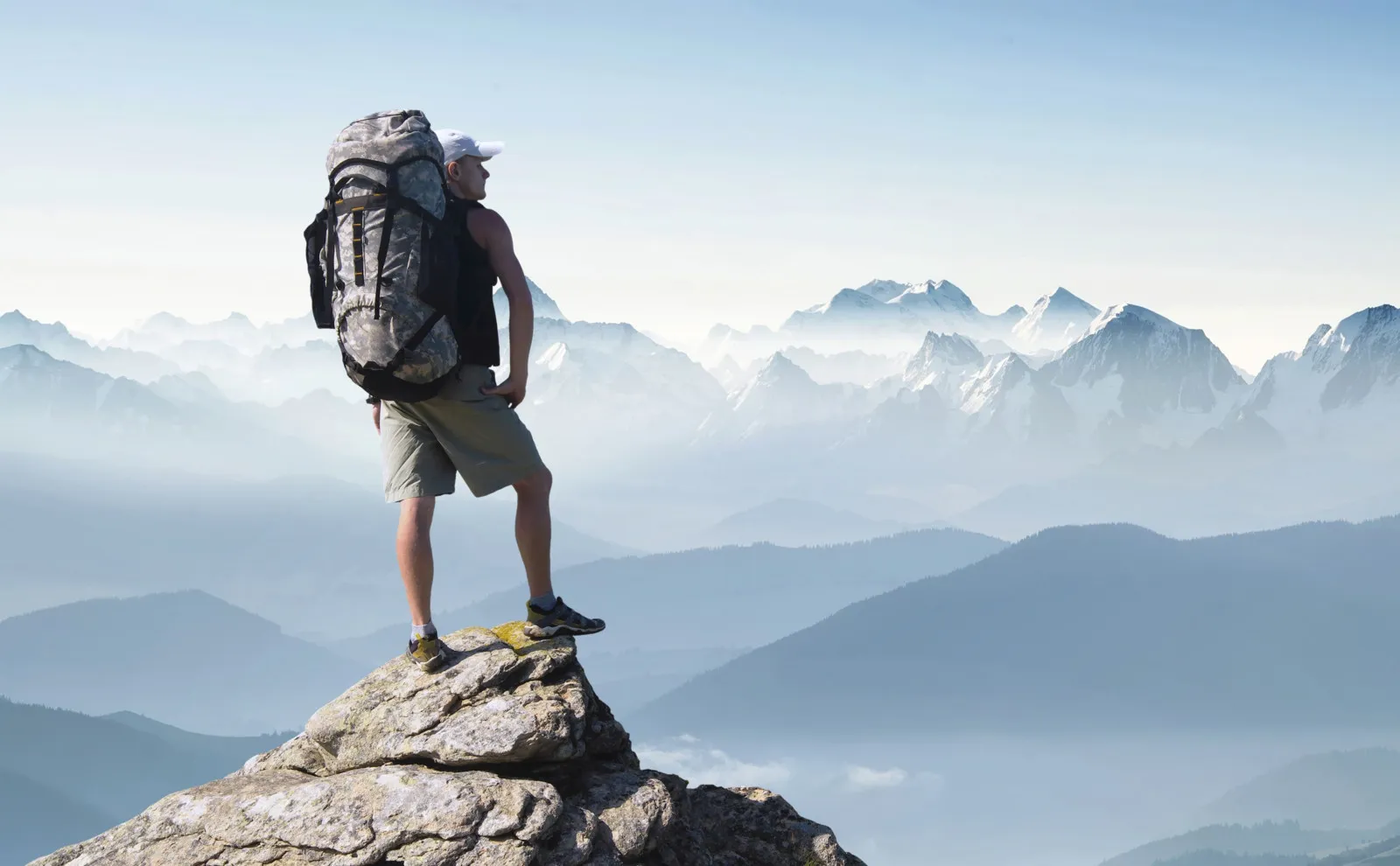 Ein Mann steht mit einem großen Rucksack auf dem Rücken auf einem Berggipfel und schaut in die Ferne.