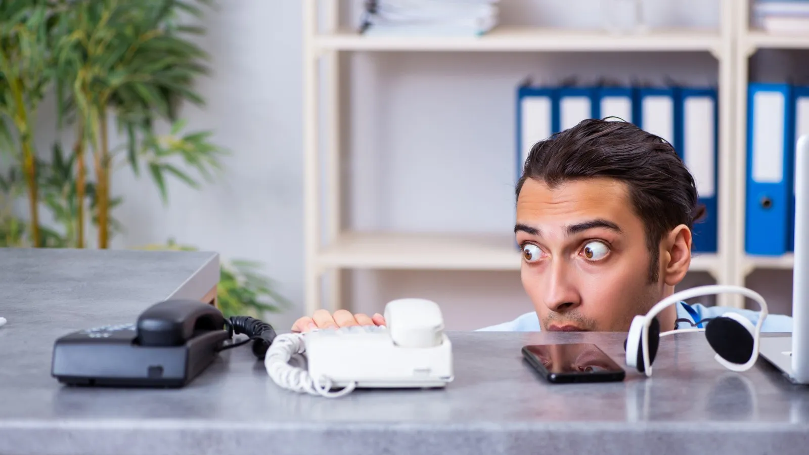 Mann versteckt sich halb hinter einem Schreibtisch und starrt verängstigt das darauf liegende weiße Telefon an.