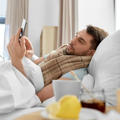kranker Mann liegt im Bett und tippt auf sein Smartphone