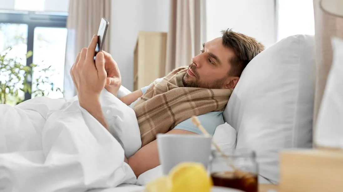 kranker Mann liegt im Bett und tippt auf sein Smartphone