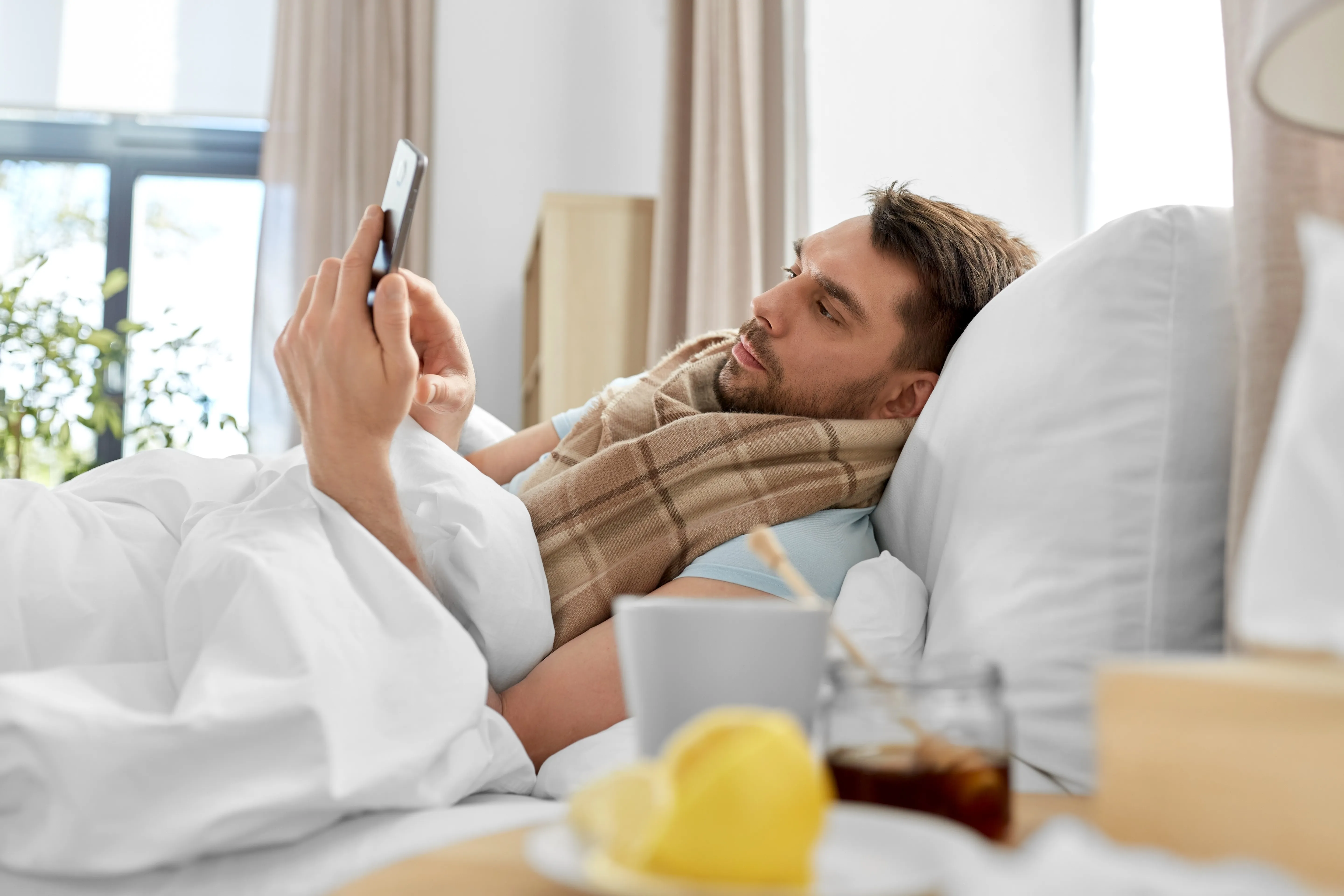 kranker Mann liegt im Bett und tippt auf sein Smartphone