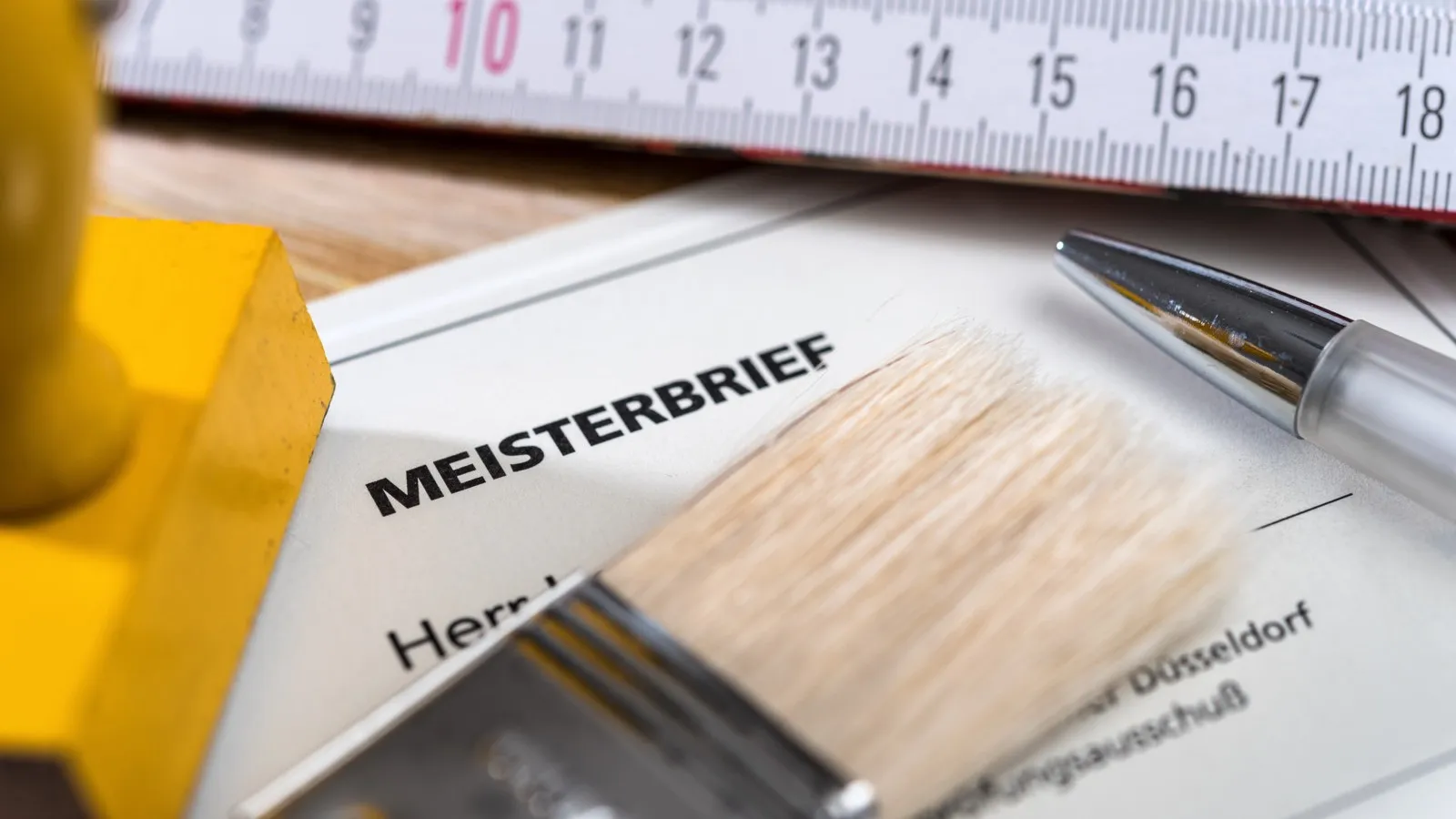 Ein Meisterbrief, auf dem ein Malerpinsel liegt, darüber liegt ein Zollstock.