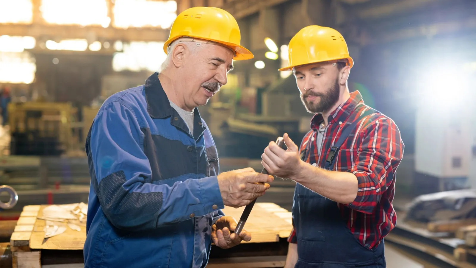 Ein junger und ein älterer Handwerker mit gelben Schutzhelmen. Sie halten einen Metallstab in den Händen und unterhalten sich darüber.