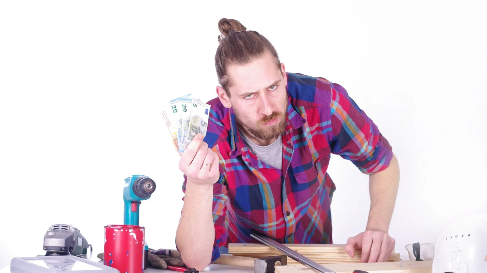 Ein Mann in einem rot-blau karierten Hemd lehnt an einem Tisch, auf dem mehrere Werkzeug verteilt sind. In der einen Hand hält er vier Euro-Banknoten hoch und schaut kritisch.