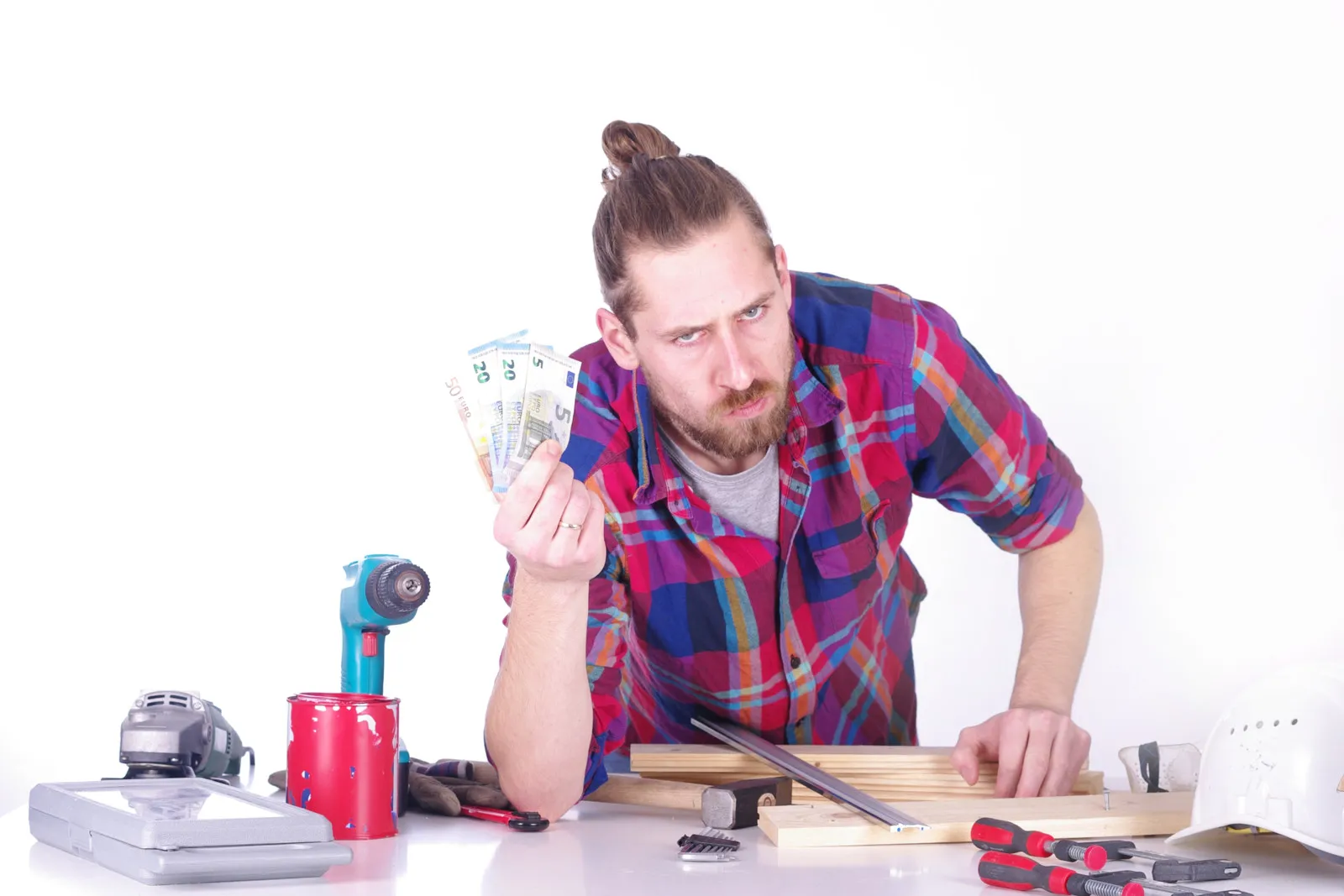 Ein Mann in einem rot-blau karierten Hemd lehnt an einem Tisch, auf dem mehrere Werkzeug verteilt sind. In der einen Hand hält er vier Euro-Banknoten hoch und schaut kritisch.