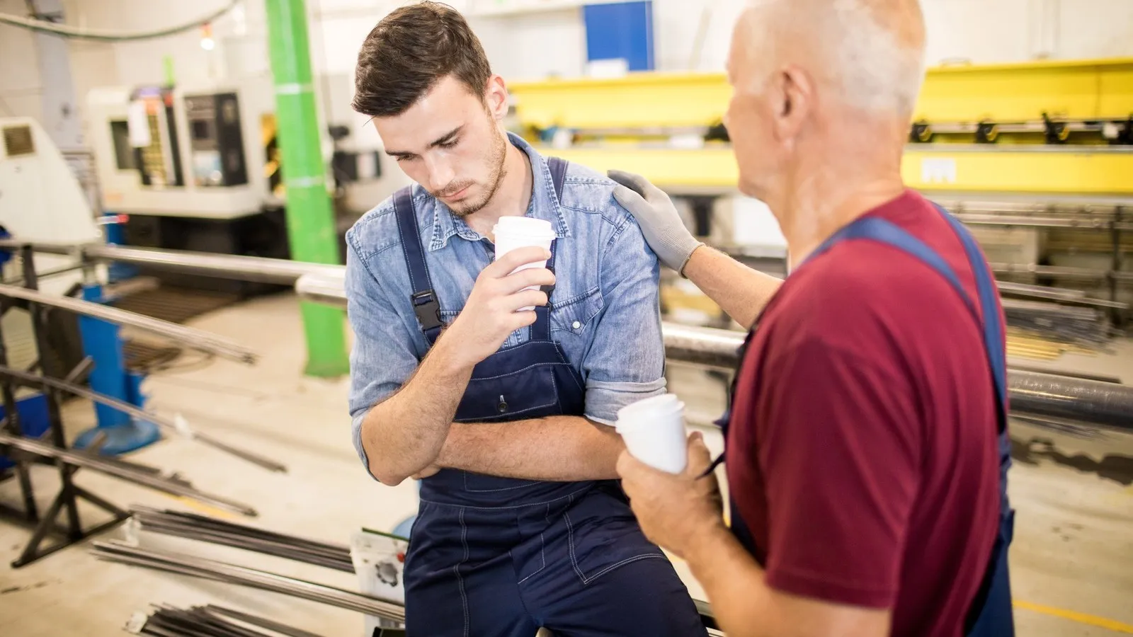 Ein junger Handwerker in einer Werkstatt sieht mit einem Kaffebecher in der Hand verunsichert zu Bode. Ein weiterer, älterer Handwerker legt ihm eine Hand auf die Schulter.