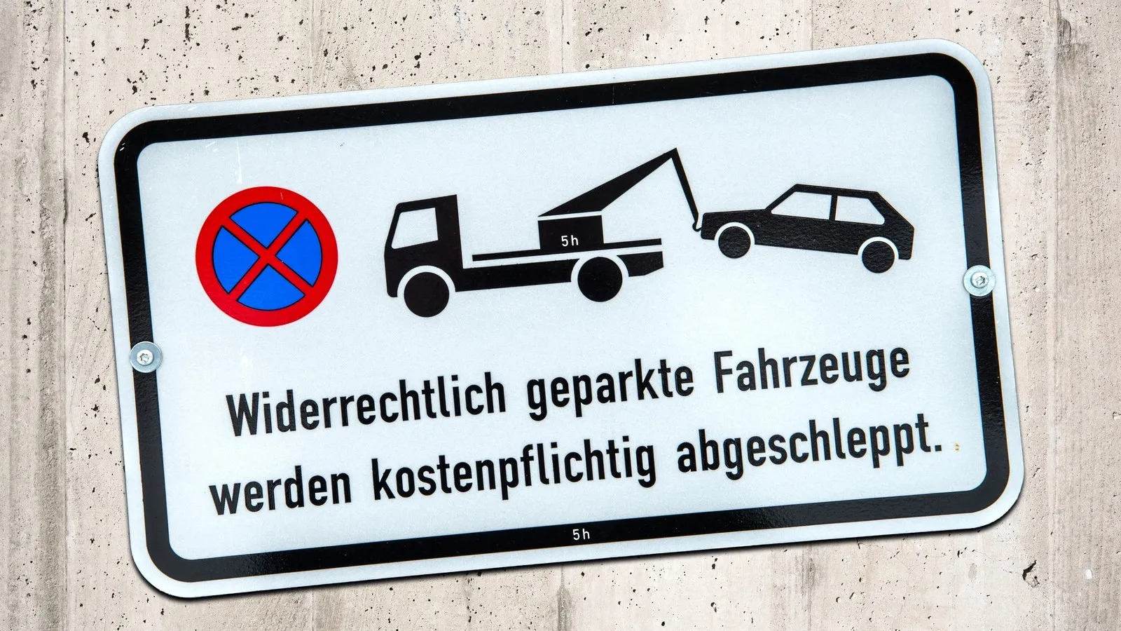Ein Parkverbotsschild mit dem Hinweis, dass "widerrechtlich geparkte Fahrzeuge kostenpflichtig abgeschleppt" werden, hängt an einer Wand.