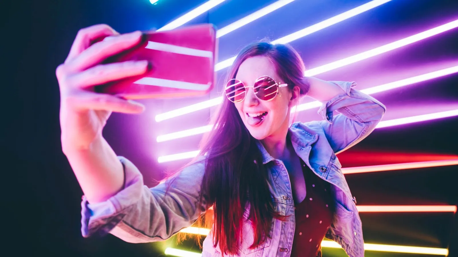 Eine Frau mit einer rosa Brille in einem beleuchteten Raum in Partyatmosphäre. In guter Laune hält sie ihr Handy für ein Selfie hoch.