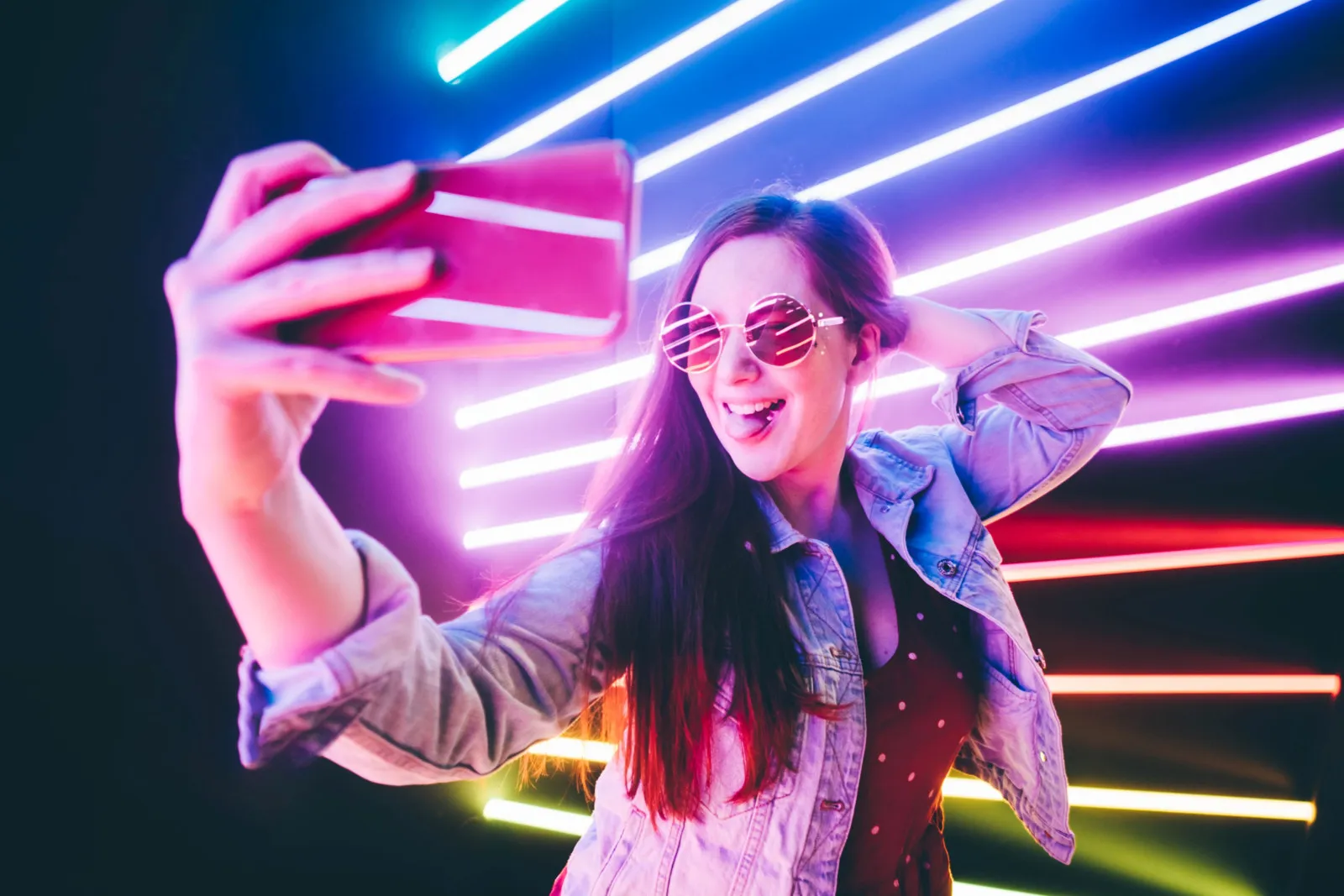 Eine Frau mit einer rosa Brille in einem beleuchteten Raum in Partyatmosphäre. In guter Laune hält sie ihr Handy für ein Selfie hoch.