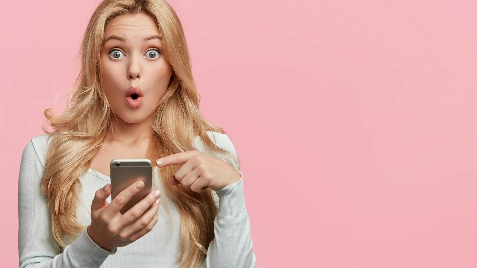 Eine blonde junge Frau schaut schockiert, zeigt auf ihr Handy, welches sie in der Hand hat. Sie steht vor einem rosafarbenen Hintergrund.
