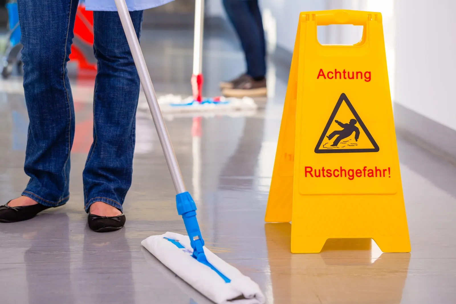 Reinigungskraft wischt einen Flur, dort ist ein gelbes Schild aufgestellt, das auf die Rutschgefahr aufmerksam macht.
