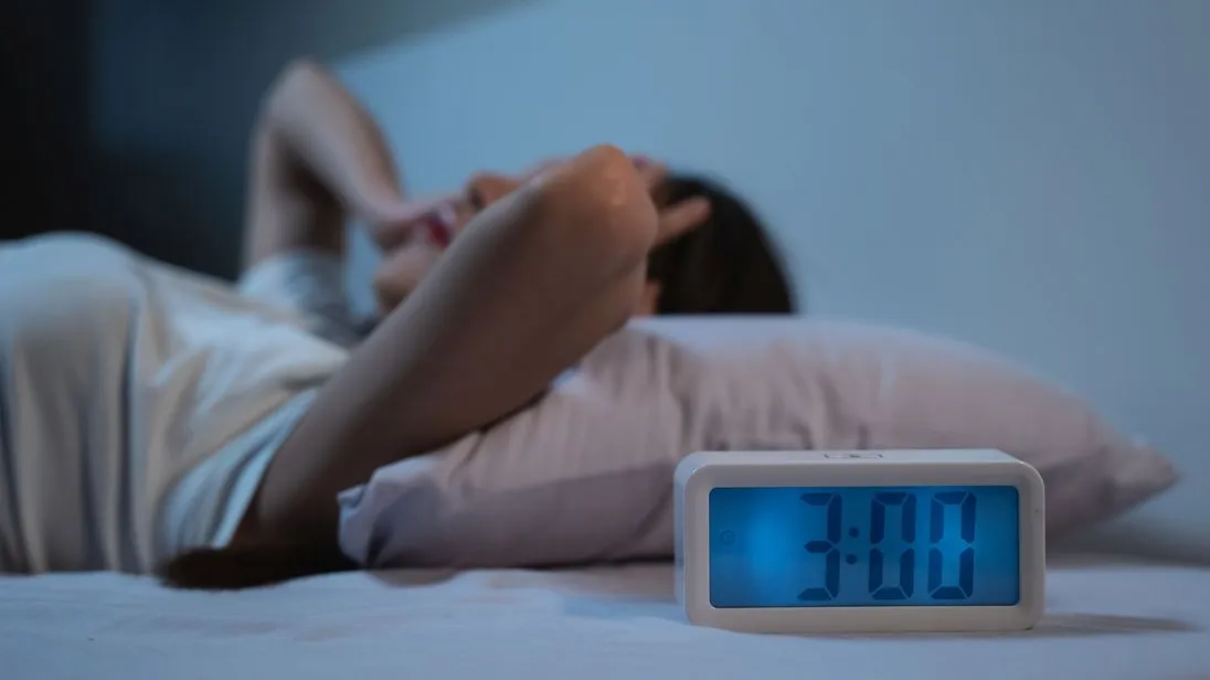 Eine Frau liegt nachts wach im Bett. Ein Wecker neben dem Bett zeigt an, dass es 3 Uhr ist.