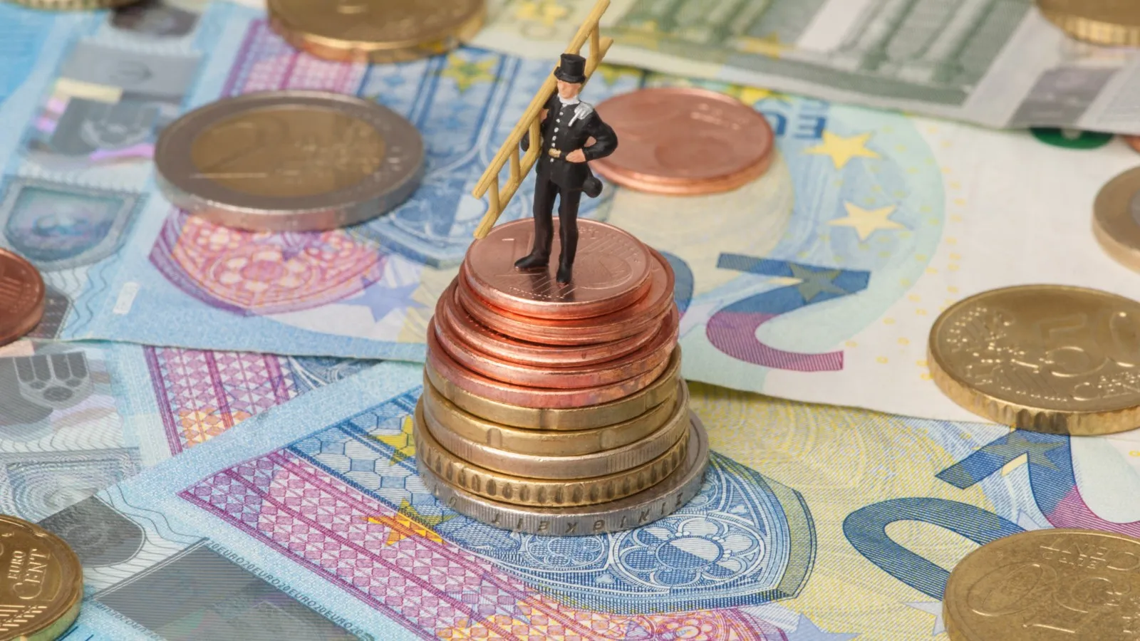 Auf mehreren 20 Euro Scheinen liegen mehrere Münzen, auf diesen Münzen steht eine Schornsteinfegerfigur.