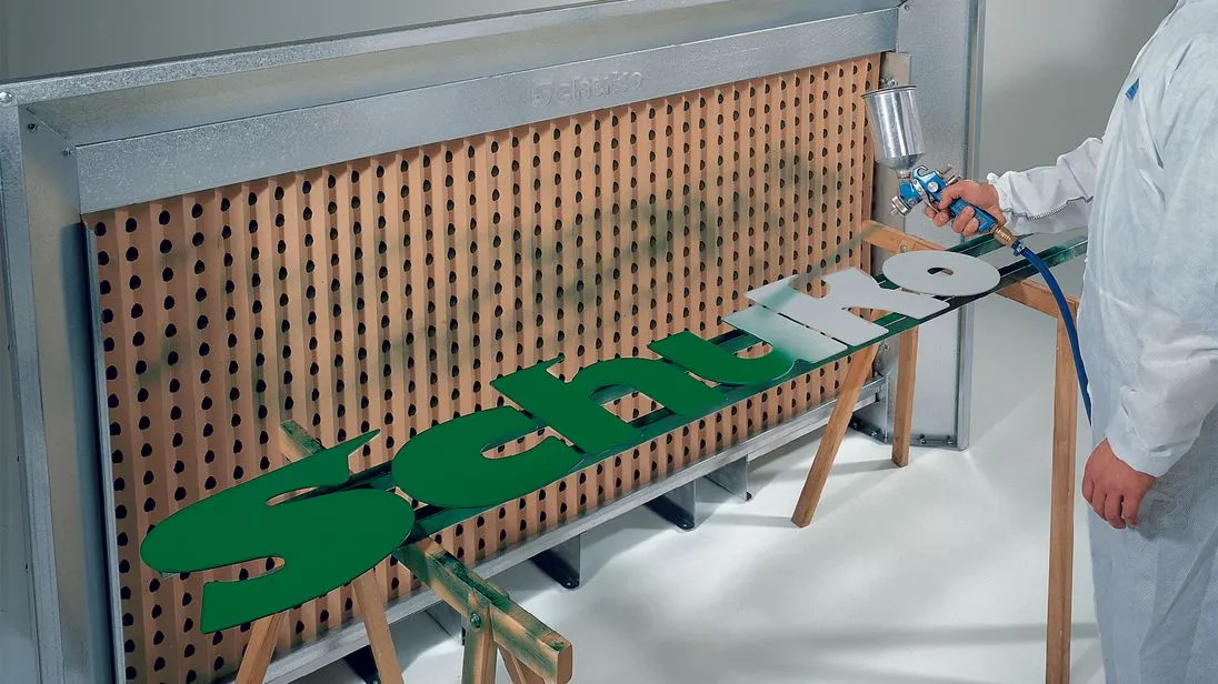 Eine Spritzwand vor der die Holzbuchstaben "Schuko" liegen. Dieser Schriftzug wird grün angesprüht. 