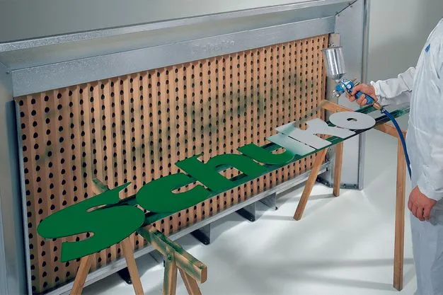 Eine Spritzwand vor der die Holzbuchstaben "Schuko" liegen. Dieser Schriftzug wird grün angesprüht. 