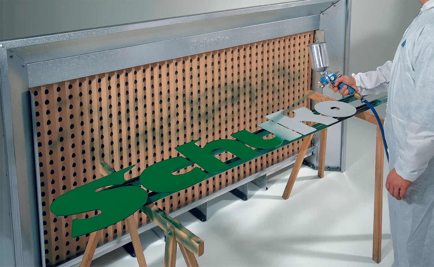 Eine Spritzwand vor der die Holzbuchstaben "Schuko" liegen. Dieser Schriftzug wird grün angesprüht. 