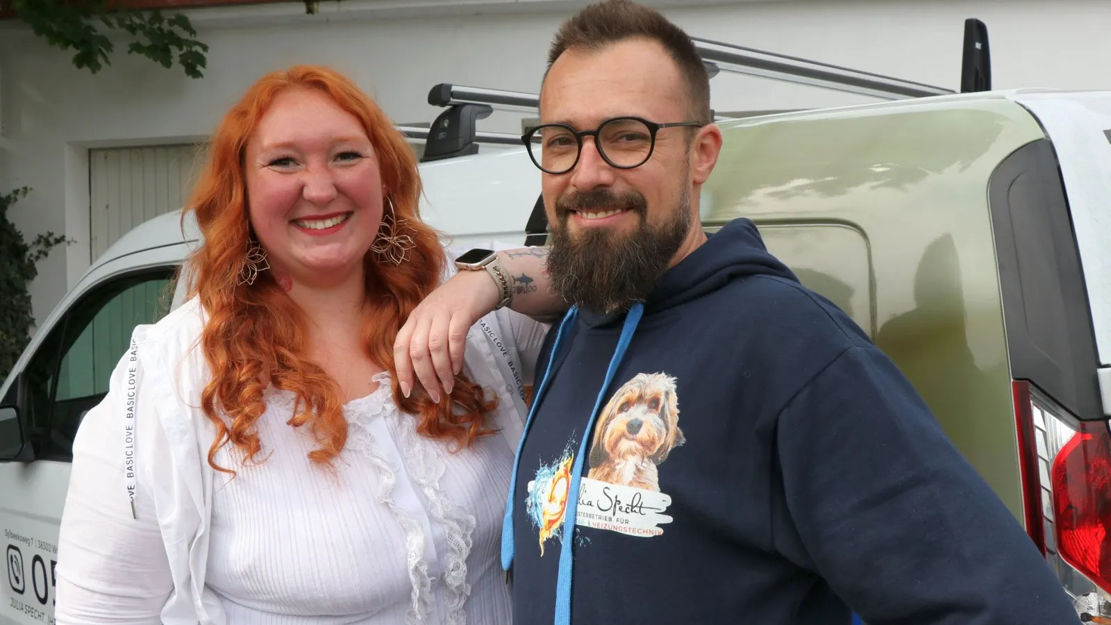 Eine rothaarige Frau und ein Mann mit Bart und Brille lächeln fürs Foto.