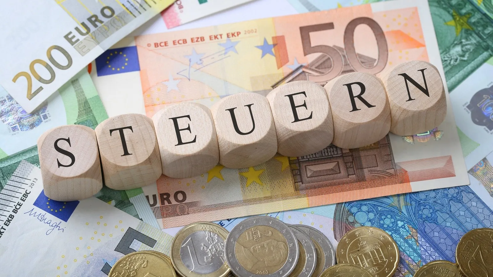 Geldscheine und Münzen liegen auf einer Oberfläche. Darauf liegen Charms aus Holz mit je einem Buchstaben, die zu dem Wort "STEUERN" angeordnet sind.
