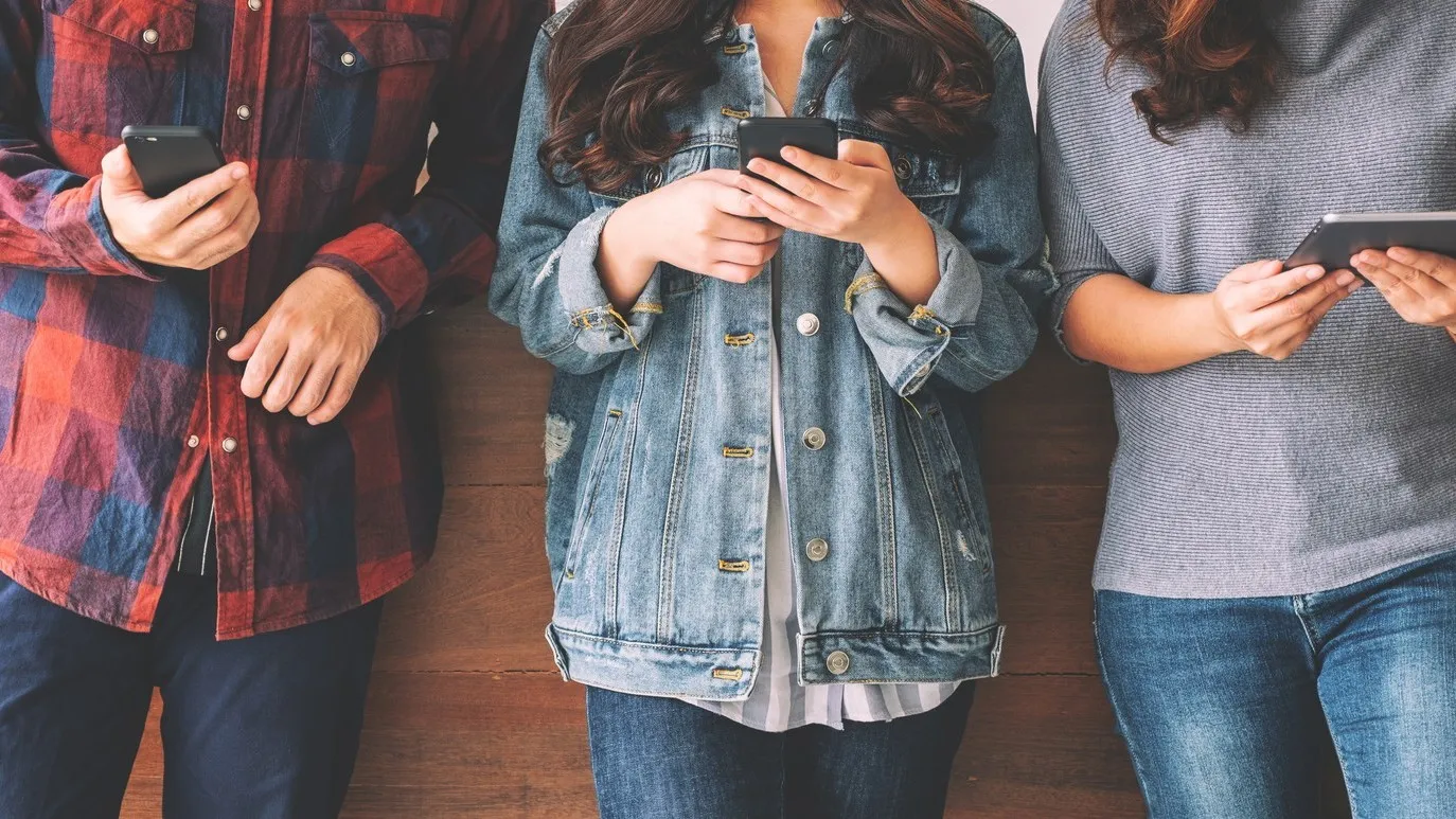 Drei junge Menschen, dessen Gesichter nicht zu sehen sind, stehen vor einer Mauer und gucken in ihr Smartphone.