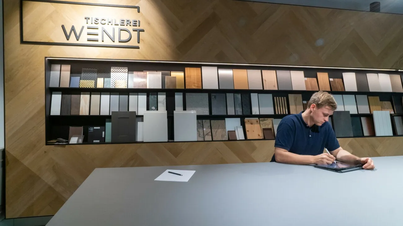 Torge Wendt an einem Tisch im Showroom. Er schreibt etwas auf. Im Hintergrund sind mehrere Muster von Materialien in einem Regal zu sehen.