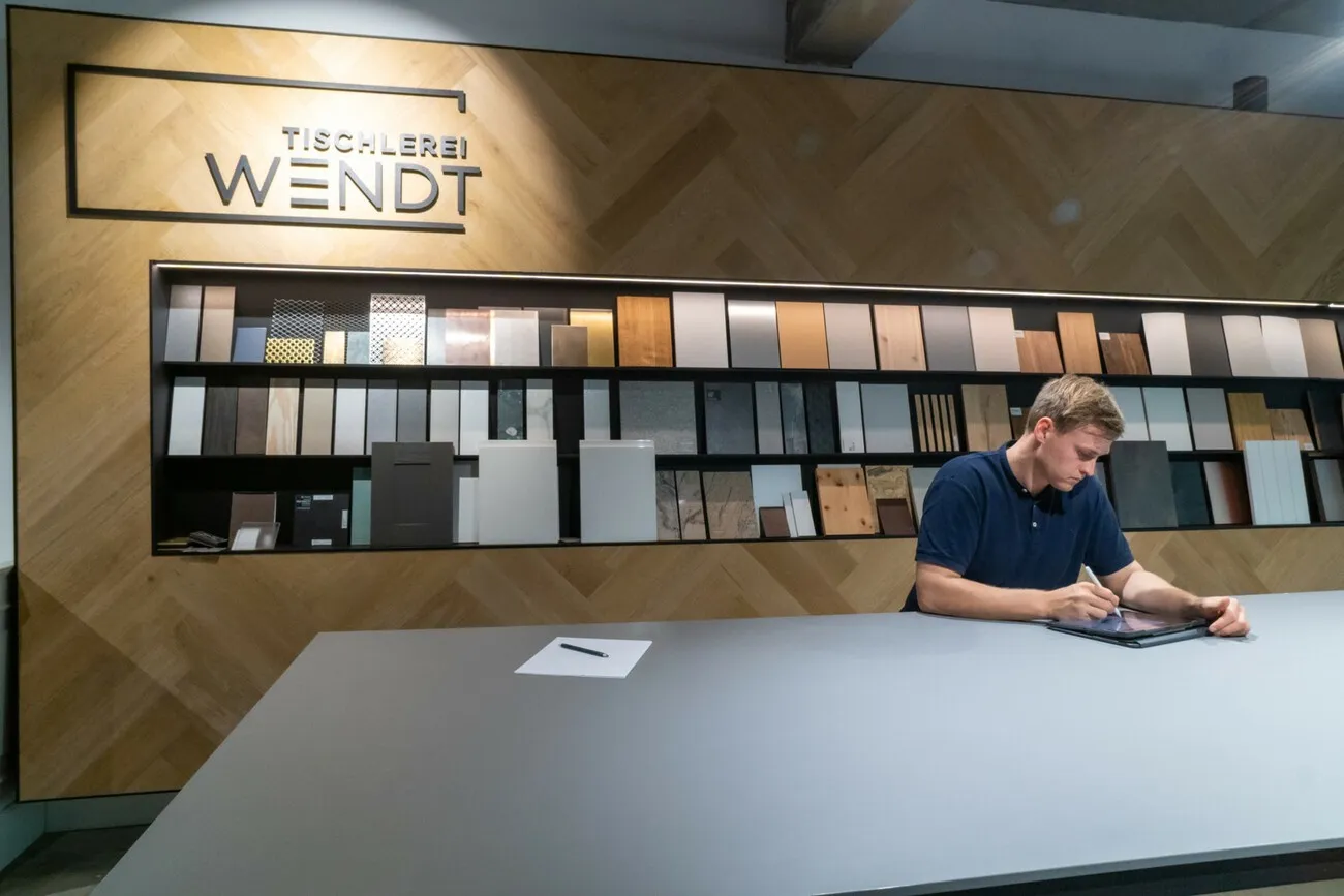 Torge Wendt an einem Tisch im Showroom. Er schreibt etwas auf. Im Hintergrund sind mehrere Muster von Materialien in einem Regal zu sehen.